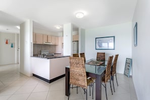 Een moderne keuken met witte kasten, een centraal eiland en een eethoek met rieten stoelen en een glazen tafel.