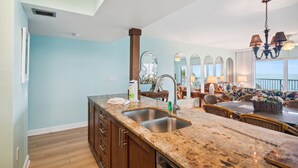 A kitchen with granite countertops, a double sink, and wooden cabinets. There is a dining area with wicker furniture and a view of the ocean.