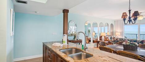 A kitchen with granite countertops, a double sink, and wooden cabinets. There is a dining area with wicker furniture and a view of the ocean.