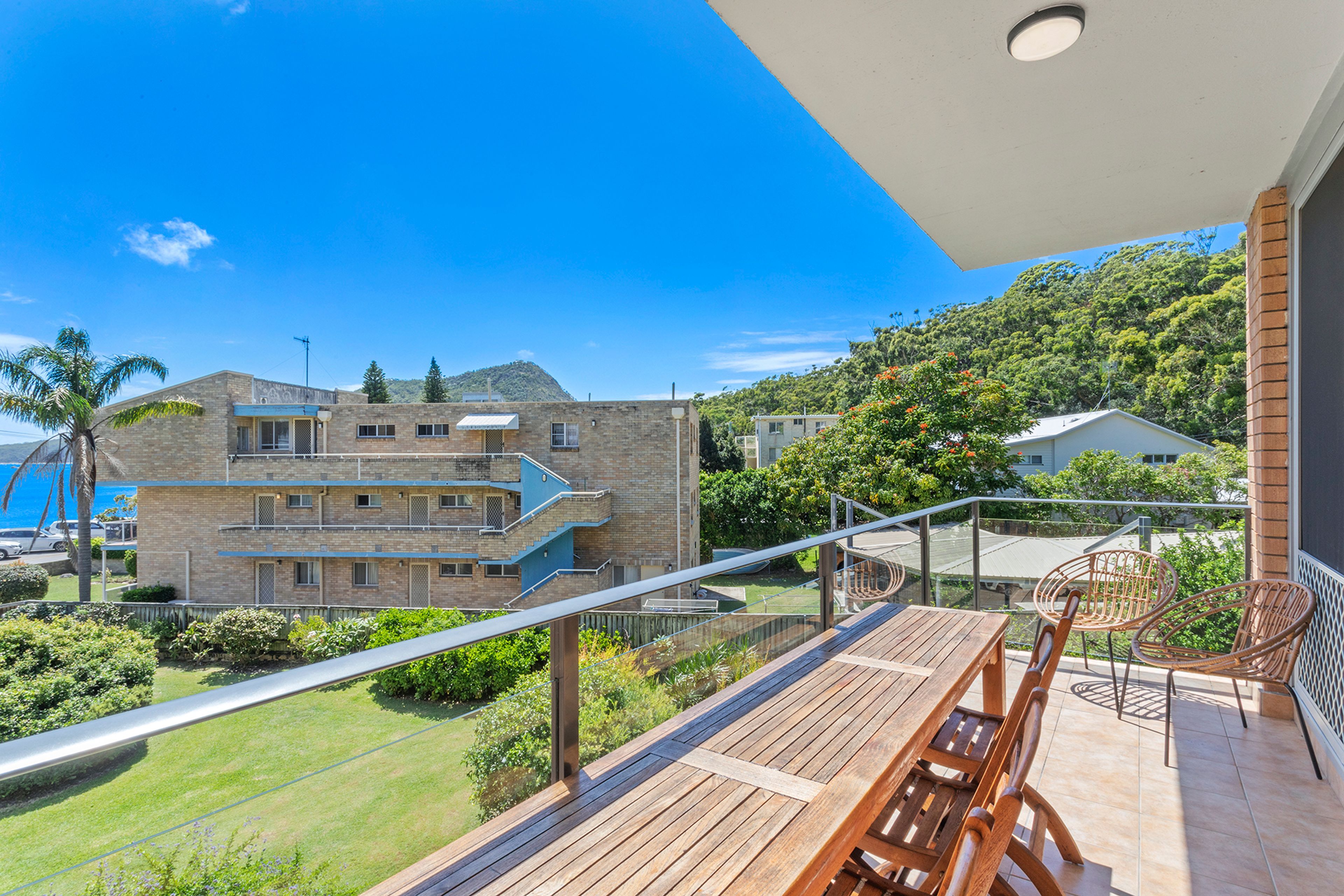 Ein Balkon mit einem Holztisch und Stühlen, der auf eine Wiese und ein Gebäude mit vielen Fenstern und einem Balkon blickt.