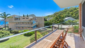 Varanda com mesa e cadeiras de madeira, com vista para área gramada e edifício com várias janelas e varanda.