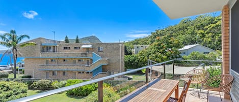 Ein Balkon mit einem Holztisch und Stühlen, der auf eine Wiese und ein Gebäude mit vielen Fenstern und einem Balkon blickt.