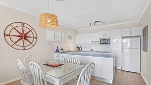 Cozinha moderna com armários brancos, uma ilha central e uma área de jantar com mesa de vidro e cadeiras brancas.