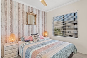 Een slaapkamer met een bed, een dressoir, een spiegel, een plafondventilator en een raam met jaloezieën.