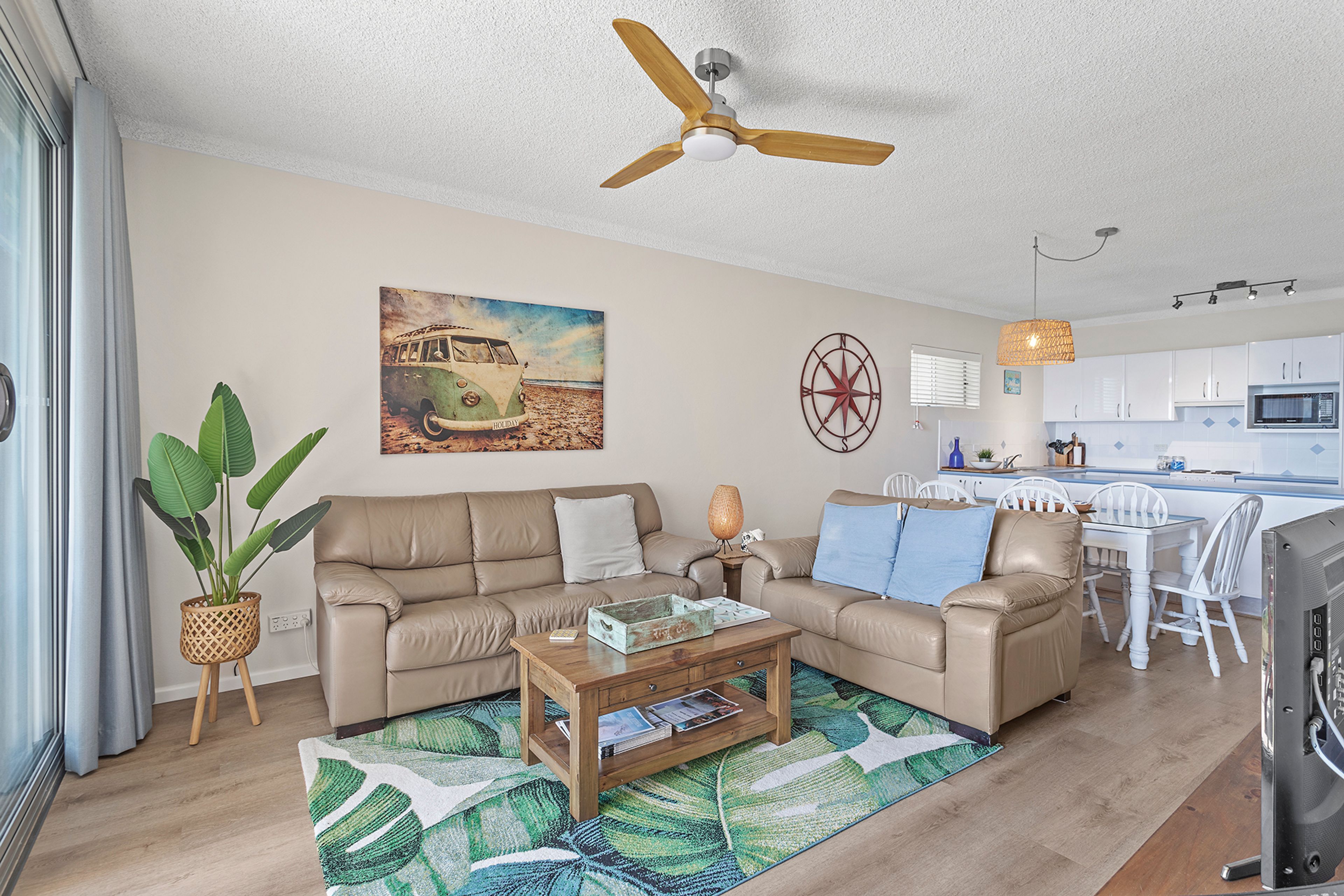 A living room with a beige sofa, a coffee table, a plant, a painting, a compass wall decoration, and a ceiling fan.