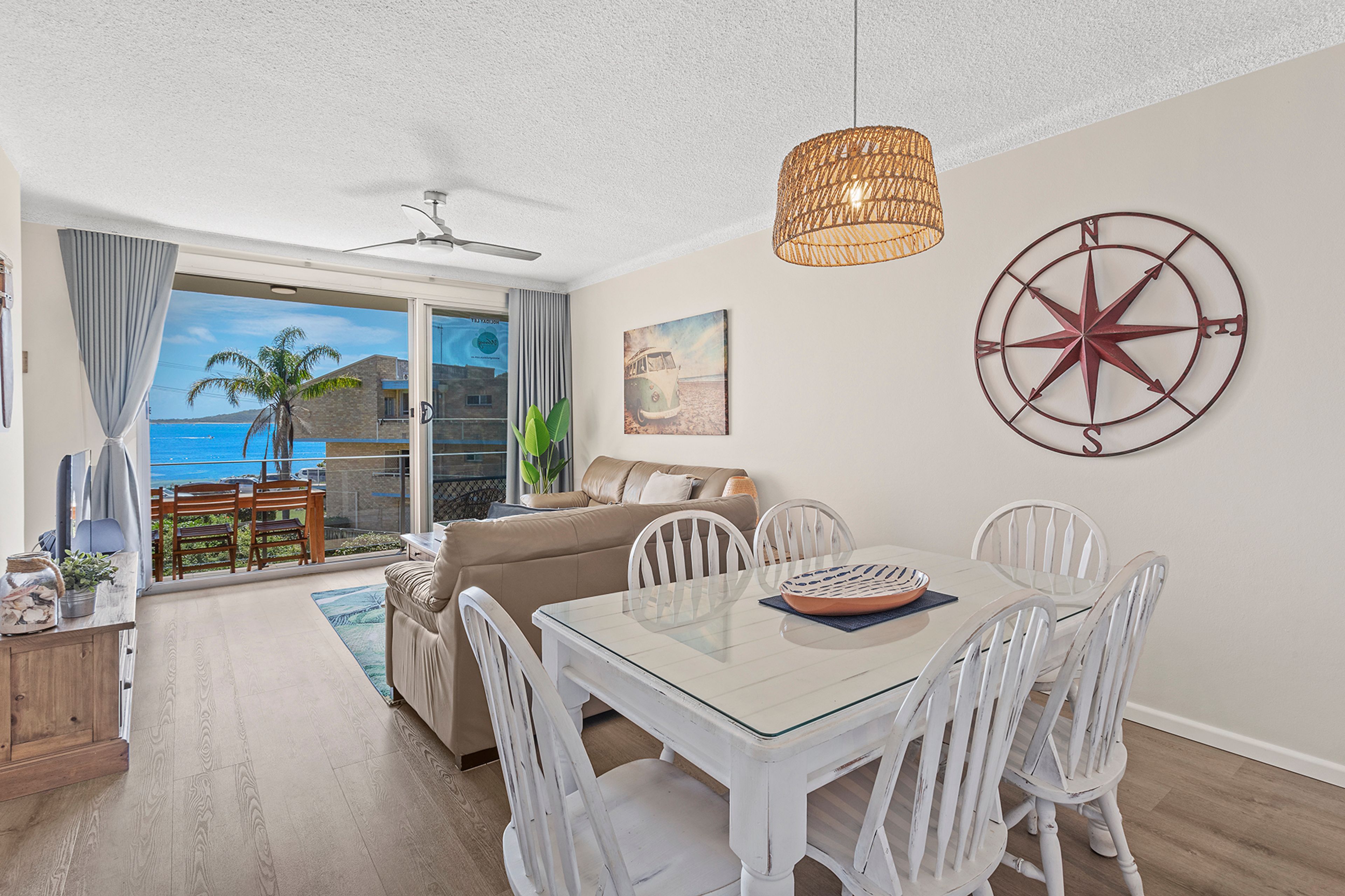 A dining area with a glass table and white chairs, a ceiling fan, and a wall-mounted compass decoration.