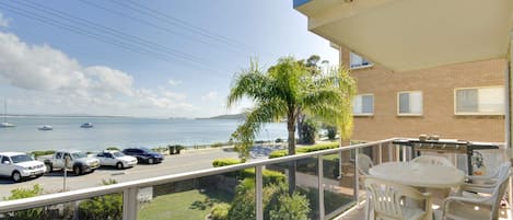 A balcony with a view of the sea, a table with chairs, and parked cars.