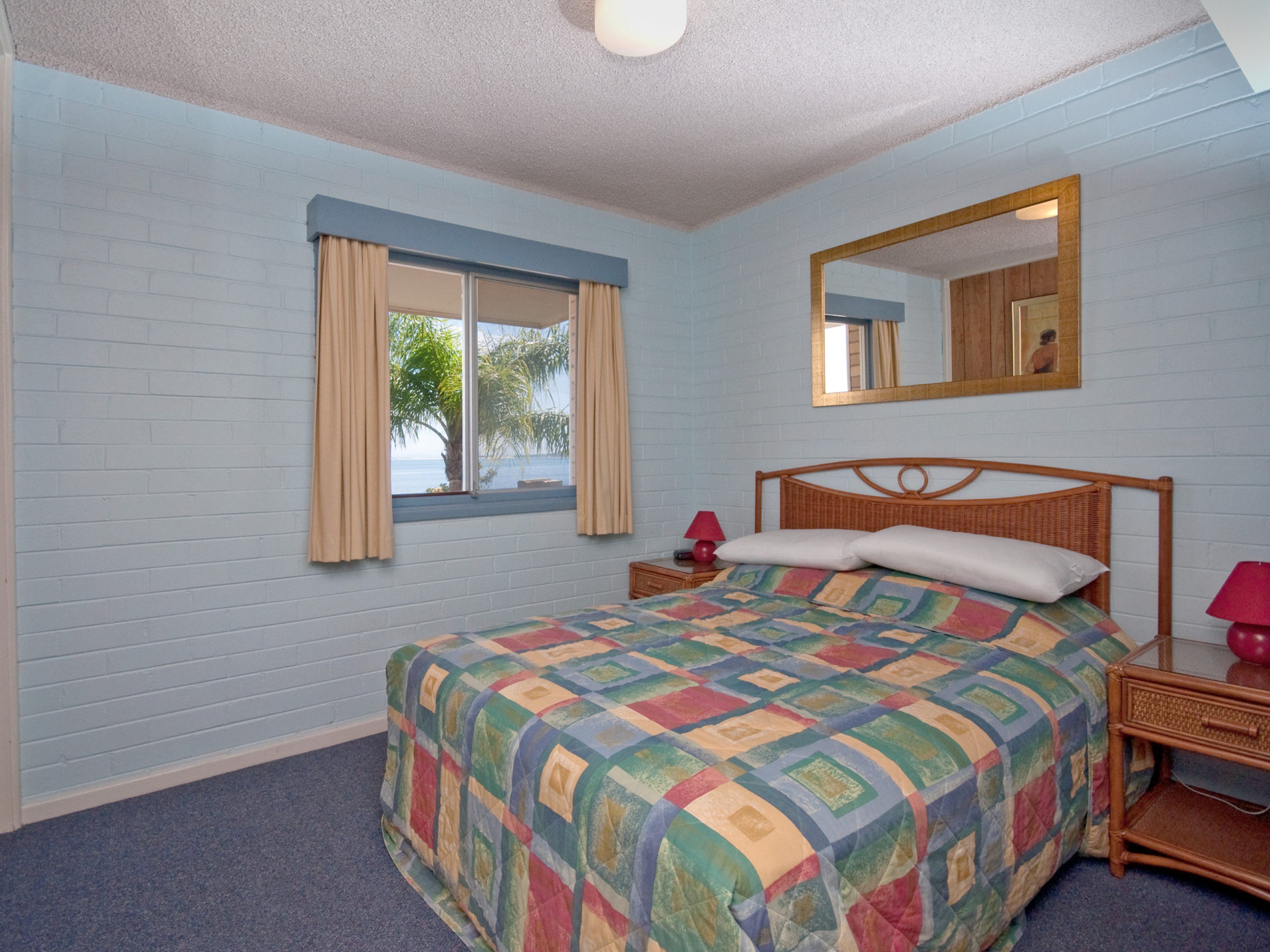 A bedroom with a bed, a window with curtains, a bedside table, a lamp, and a mirror on the wall.