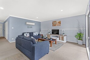 A modern living room with a grey sofa, a coffee table, a TV, and decorative plants. - Horizons Getaway at the Bay (Salamander Bay)