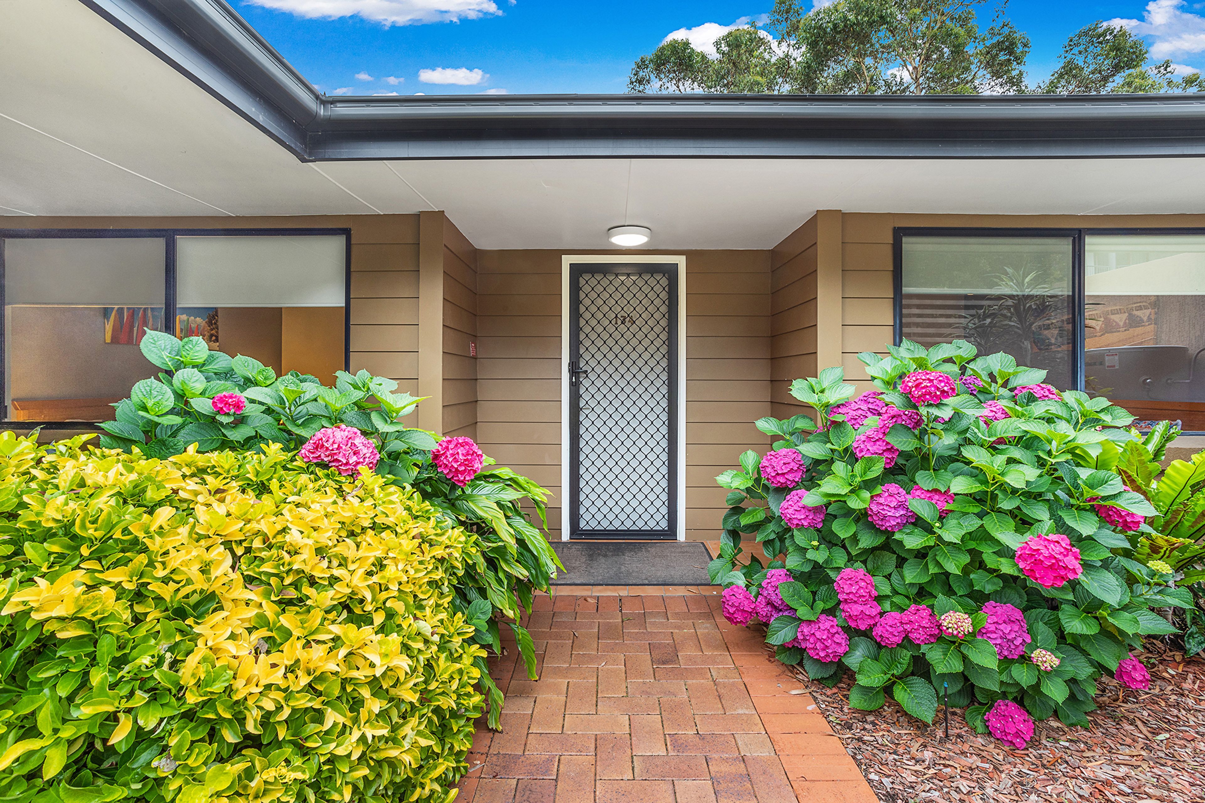 A modern building entrance with a clear sky, a metal gate, and well-maintained hedges with pink flowers.