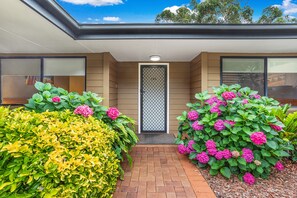 A modern building entrance with a clear sky, a metal gate, and well-maintained hedges with pink flowers. - Horizons Getaway at the Bay (Salamander Bay)