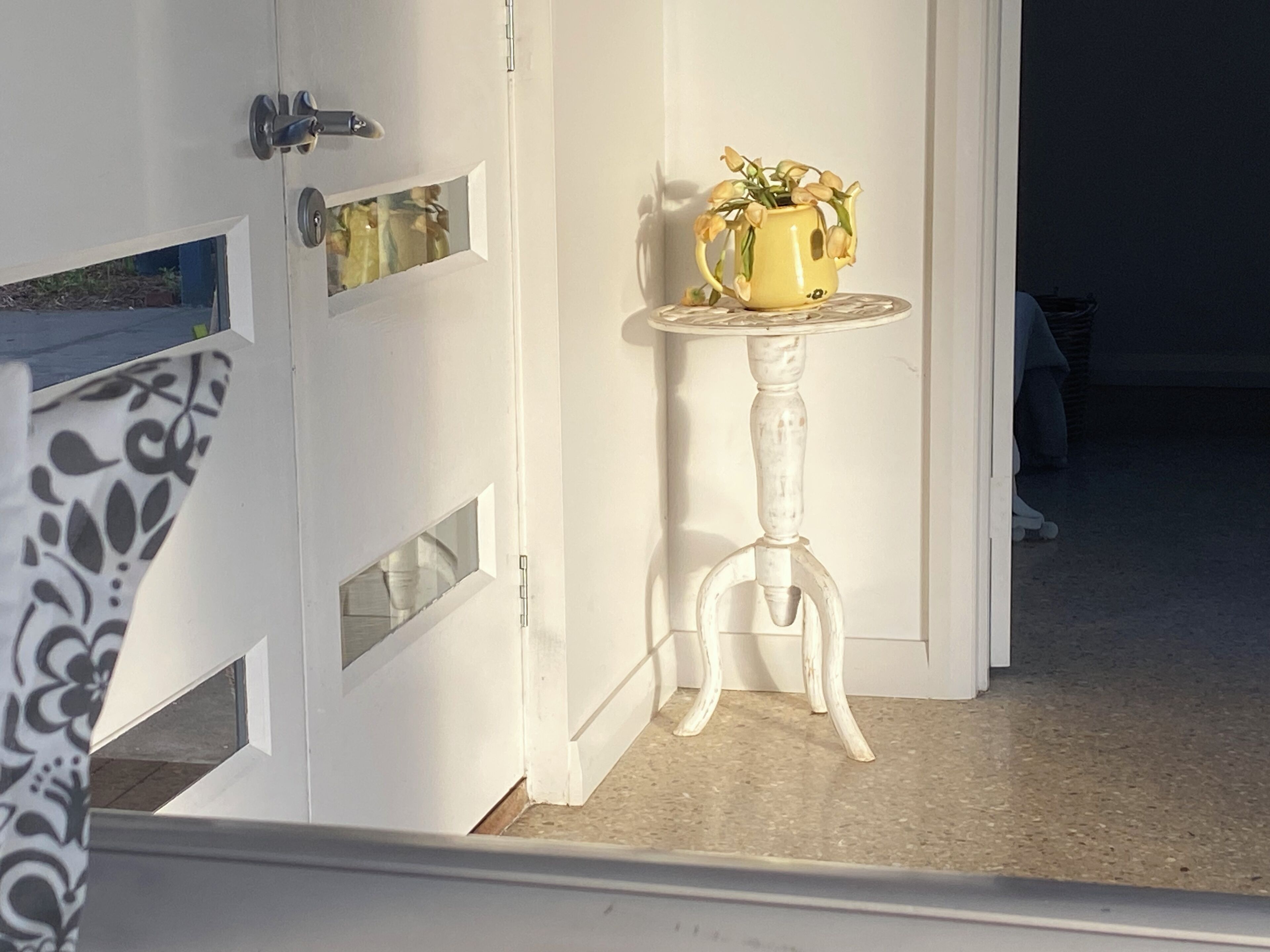 Eine weiße Tür mit einem kleinen Fenster, ein weißer Tisch mit einer gelben Vase und ein teilweise sichtbarer, gemusterter Vorhang.