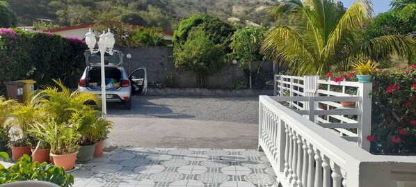 Un camino de entrada cerrado por una reja conduce a un coche estacionado con las puertas abiertas, rodeado de plantas en macetas y una valla decorativa.