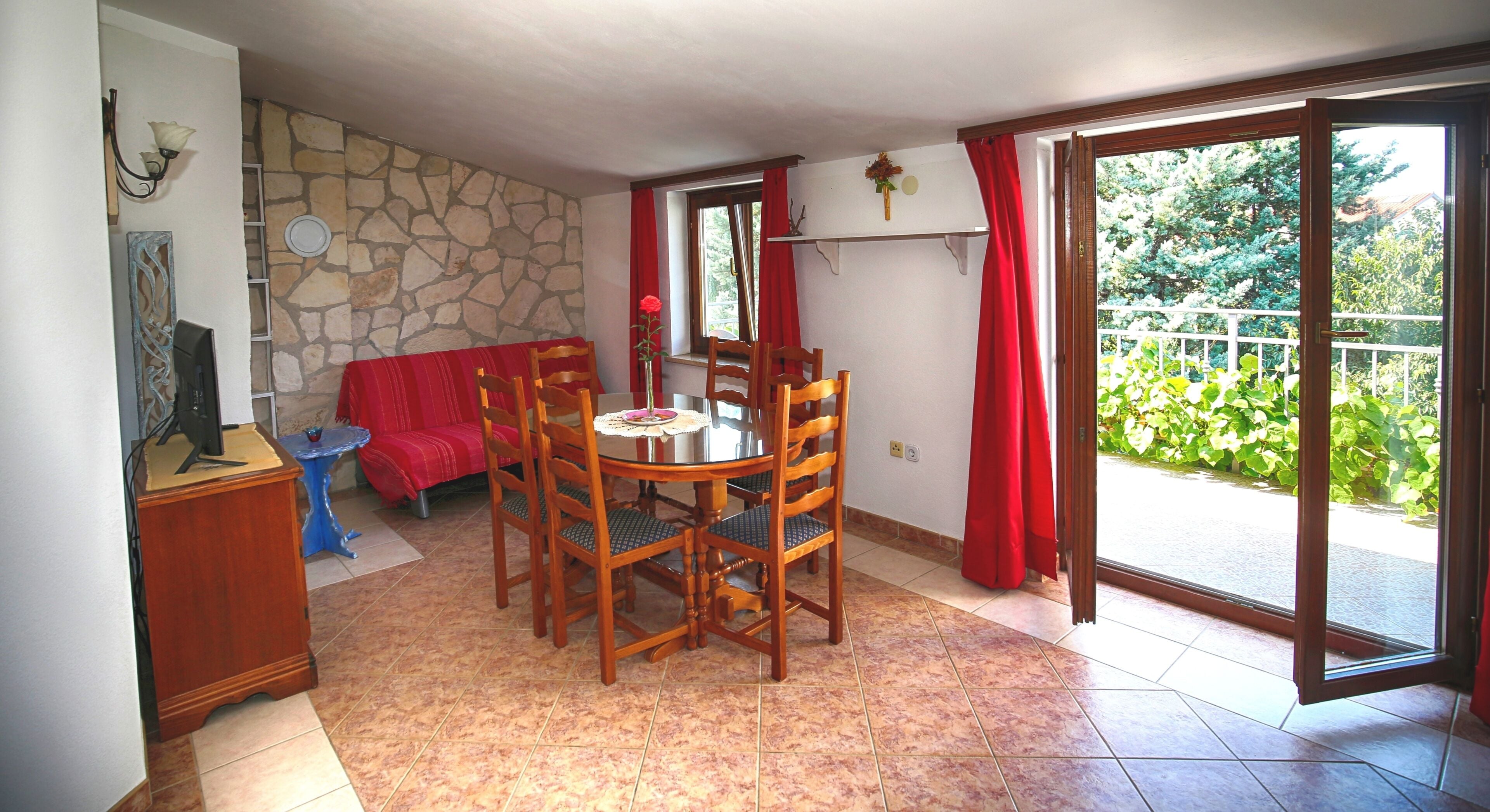 A dining area with a round table and chairs, a red sofa, a stone wall, and a view through a glass door.