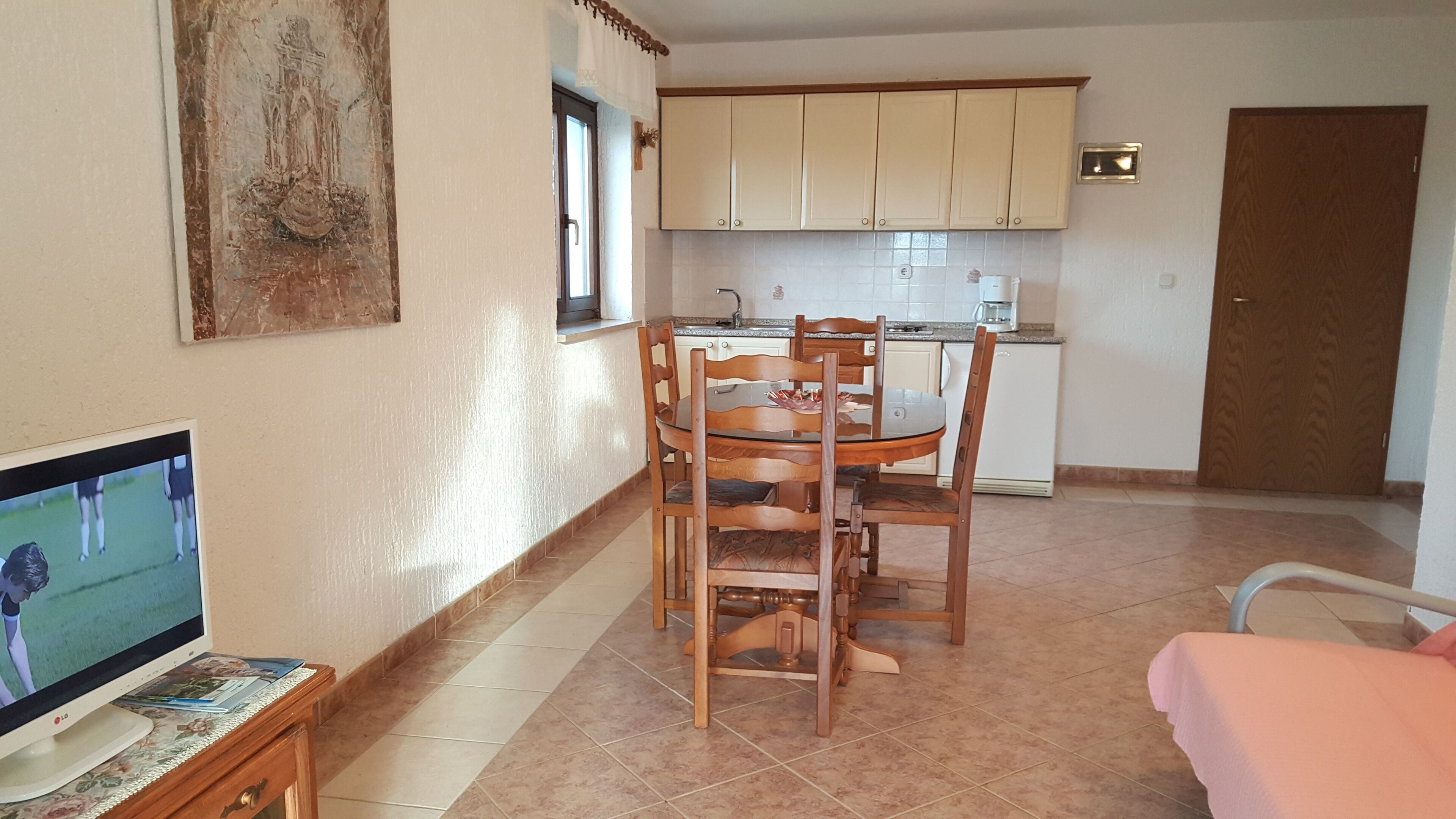 Una cocina funcional con armarios blancos, una mesa de comedor con sillas, un televisor sobre un mueble de madera y una puerta al fondo.