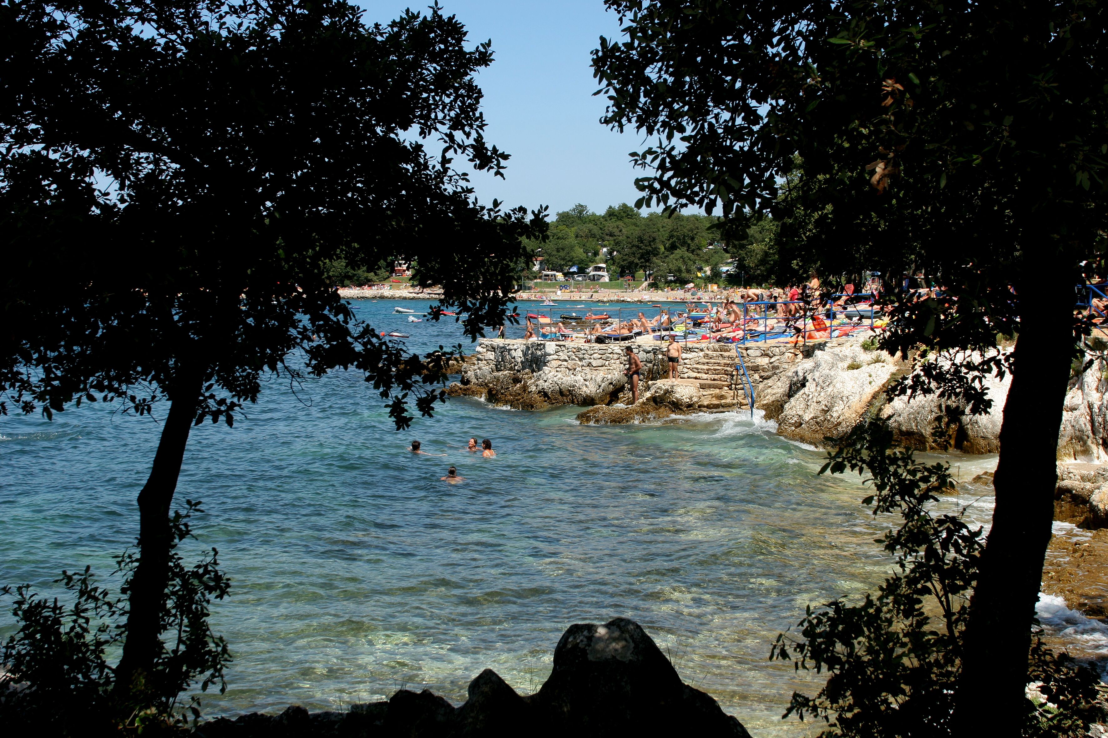 Ein Strand mit klarem Wasser, felsigem Ufer und badenden Personen.