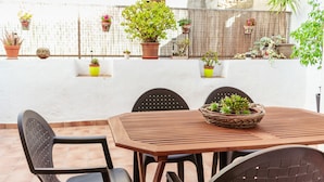 Une terrasse avec une table et des chaises en bois, entourée de plantes en pot et d’un mur blanc.
