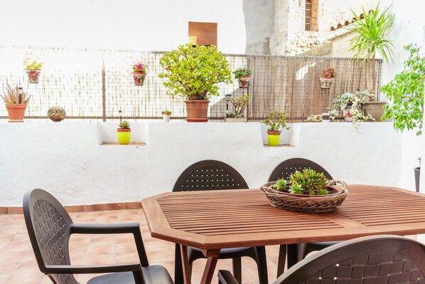 A patio with a wooden table and chairs, surrounded by potted plants and a white wall.