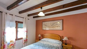 Une chambre à coucher comprenant un lit, deux tables de chevet, une fenêtre avec des rideaux, un plafond avec des poutres apparentes et un tableau accroché au mur.