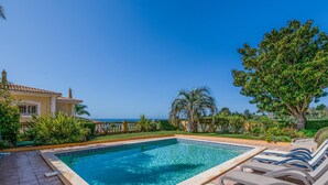 A rectangular swimming pool with clear blue water, surrounded by a tiled deck. There are lounge chairs with metal frames and cushioned seats. A large tree and various shrubs are visible in the background. - Casa Mira Luz, villa with private pool (Praia da Luz)