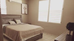 A bedroom with a bed, a nightstand, a potted plant, and a window. - Rent It (Aguadilla)