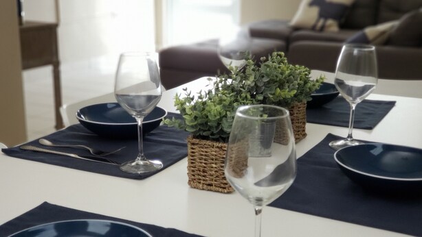 A dining table set with blue plates, wine glasses, and silverware, accompanied by a small potted plant. - Rent It (Aguadilla)
