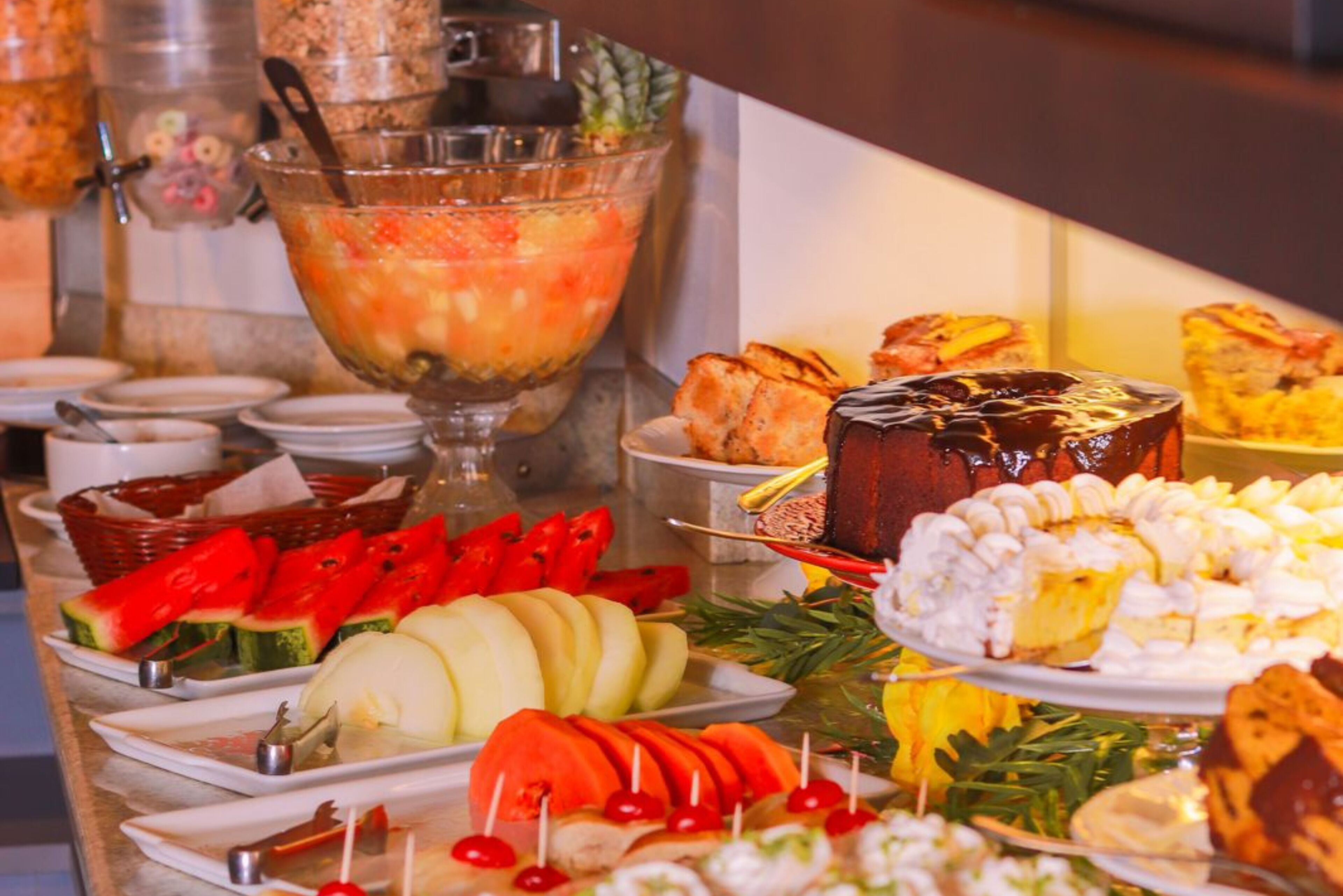 A buffet with various food items including fruits, pastries, and a large bowl of fruit salad.