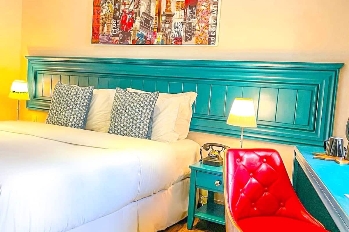 A bed with white bedding, a teal headboard, and patterned pillows. There is a red chair and a small table with a lamp.