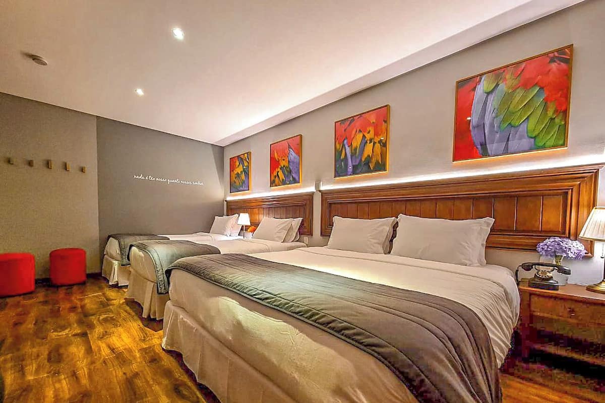A hotel room with three beds, a wooden headboard, a red ottoman, and a wall with abstract paintings.