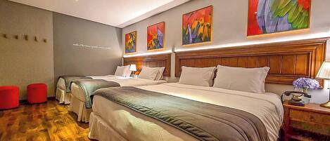 A hotel room with three beds, a wooden headboard, a red ottoman, and a wall with abstract paintings.