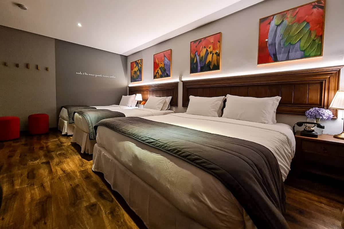 A hotel room with three beds, wooden flooring, a headboard with built-in lighting, and artwork on the walls.