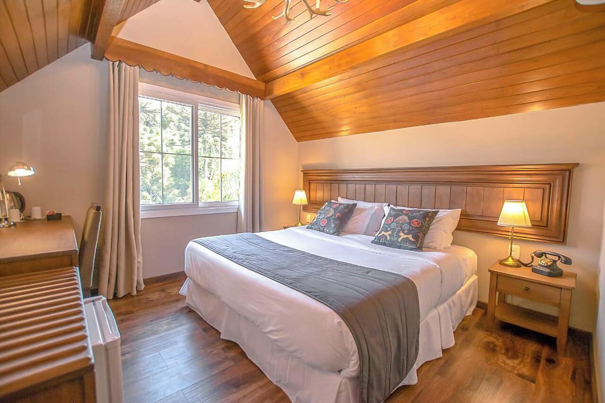 A bedroom with a wooden ceiling, a large bed with multiple pillows, a bedside table with a lamp, and a window with curtains.