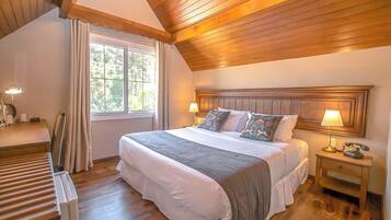 A bedroom with a wooden ceiling, a large bed with multiple pillows, a bedside table with a lamp, and a window with curtains.