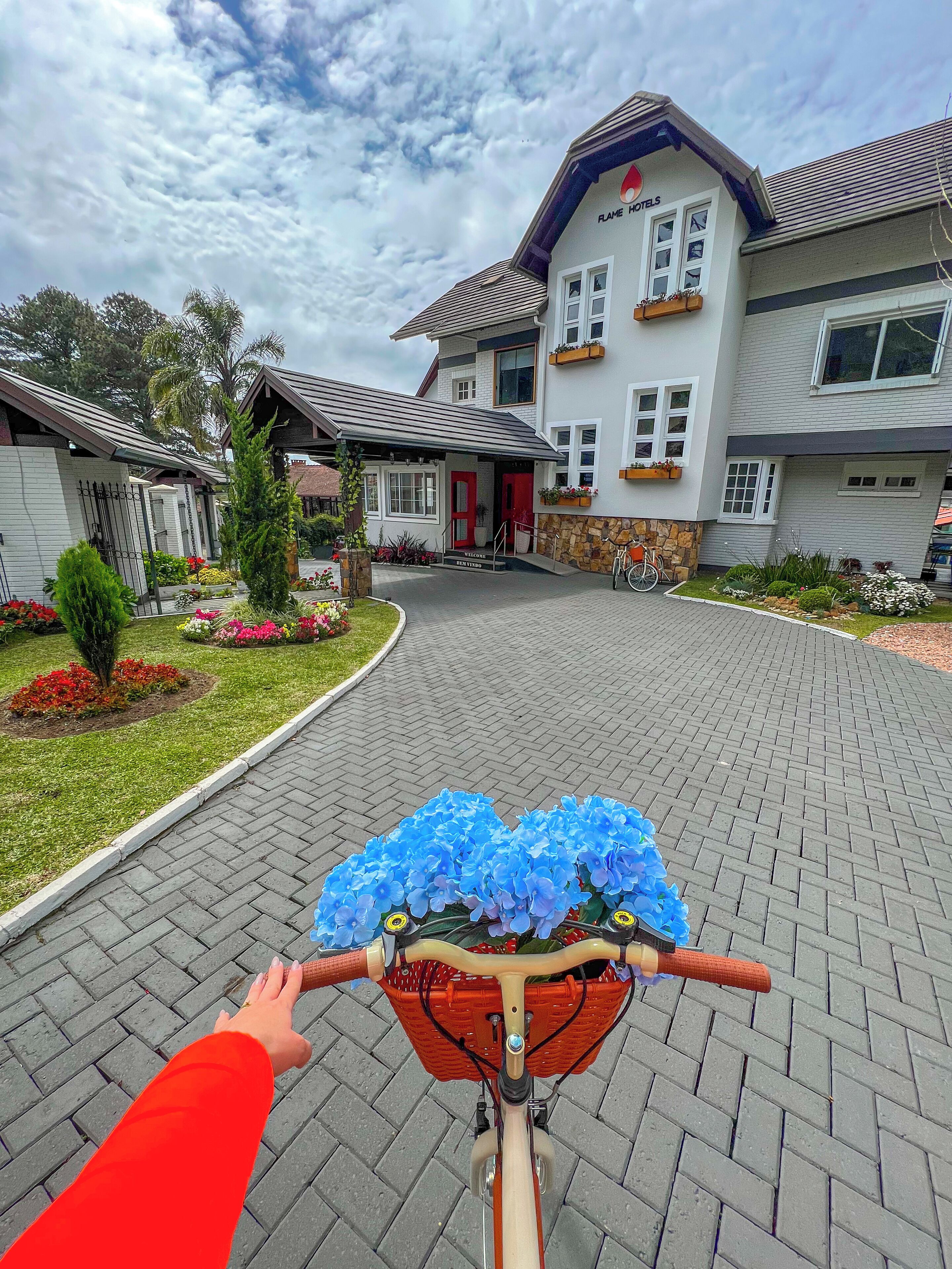 A bicycle with a basket containing blue flowers, approaching a hotel entrance with a red door and a sign that reads "Welcome."
