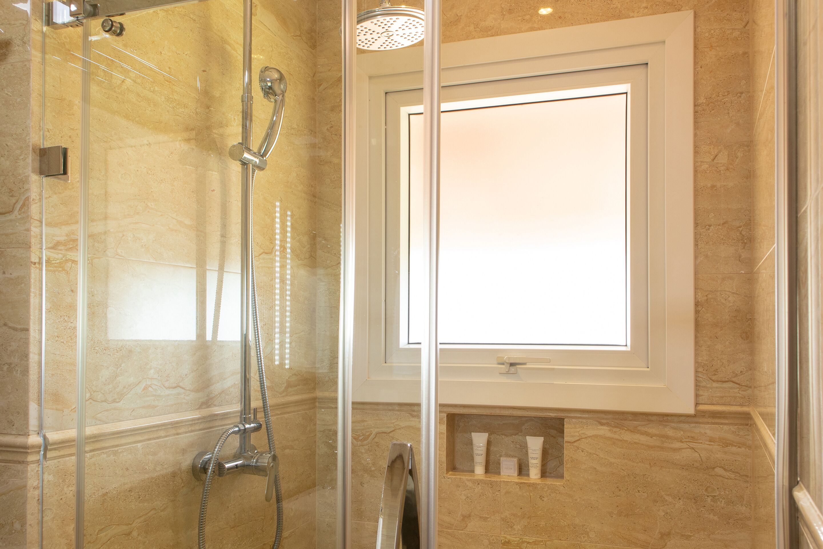Un baño con ducha envuelta en cristal, pared de mármol y estante empotrado con artículos de aseo.