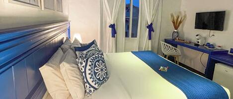 A bedroom with a bed, a desk, a chair, a TV, and a window with curtains.
