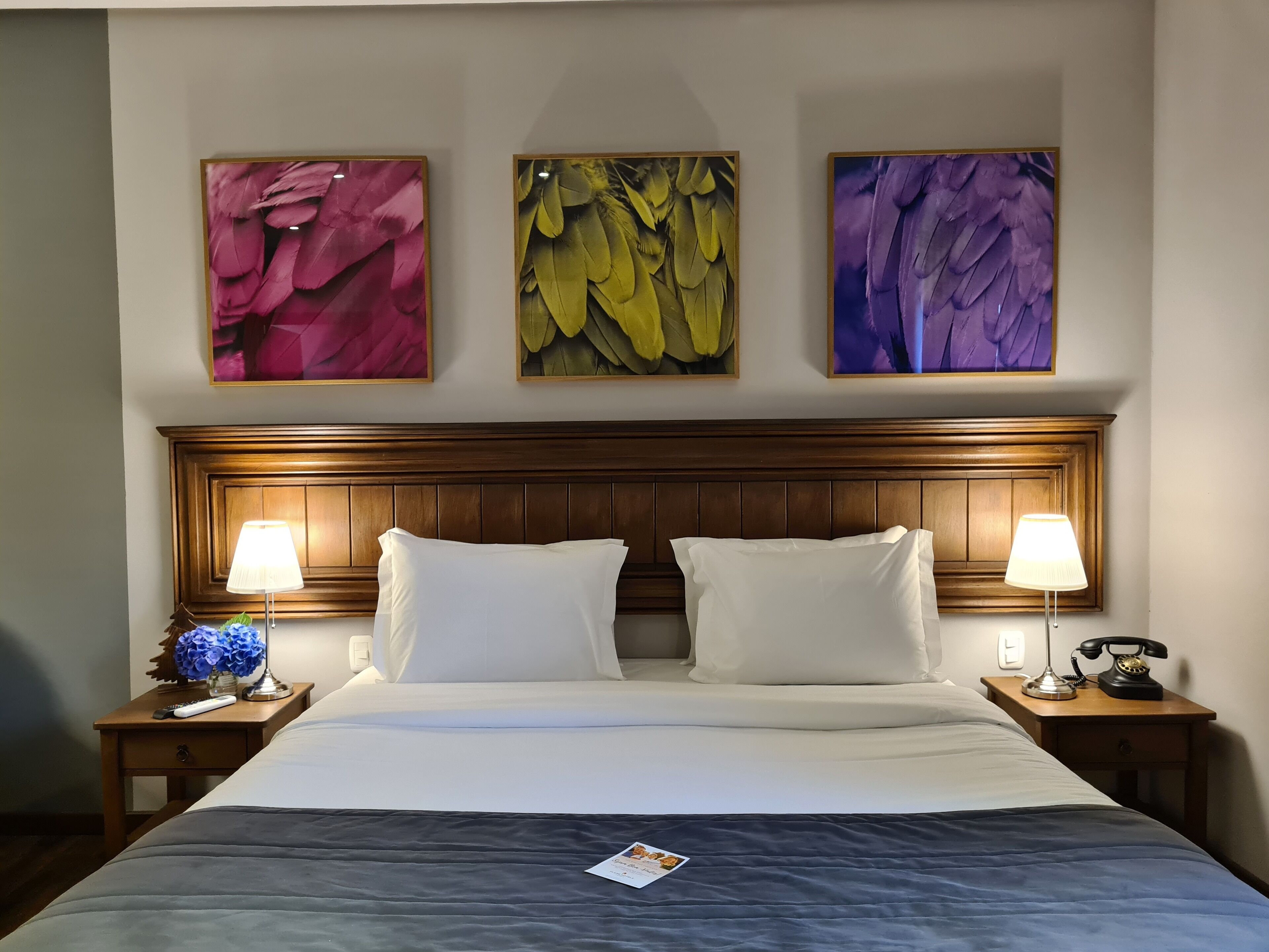 A hotel room with a wooden headboard, two bedside lamps, a remote control, a telephone, and a bedside table with a vase of flowers.