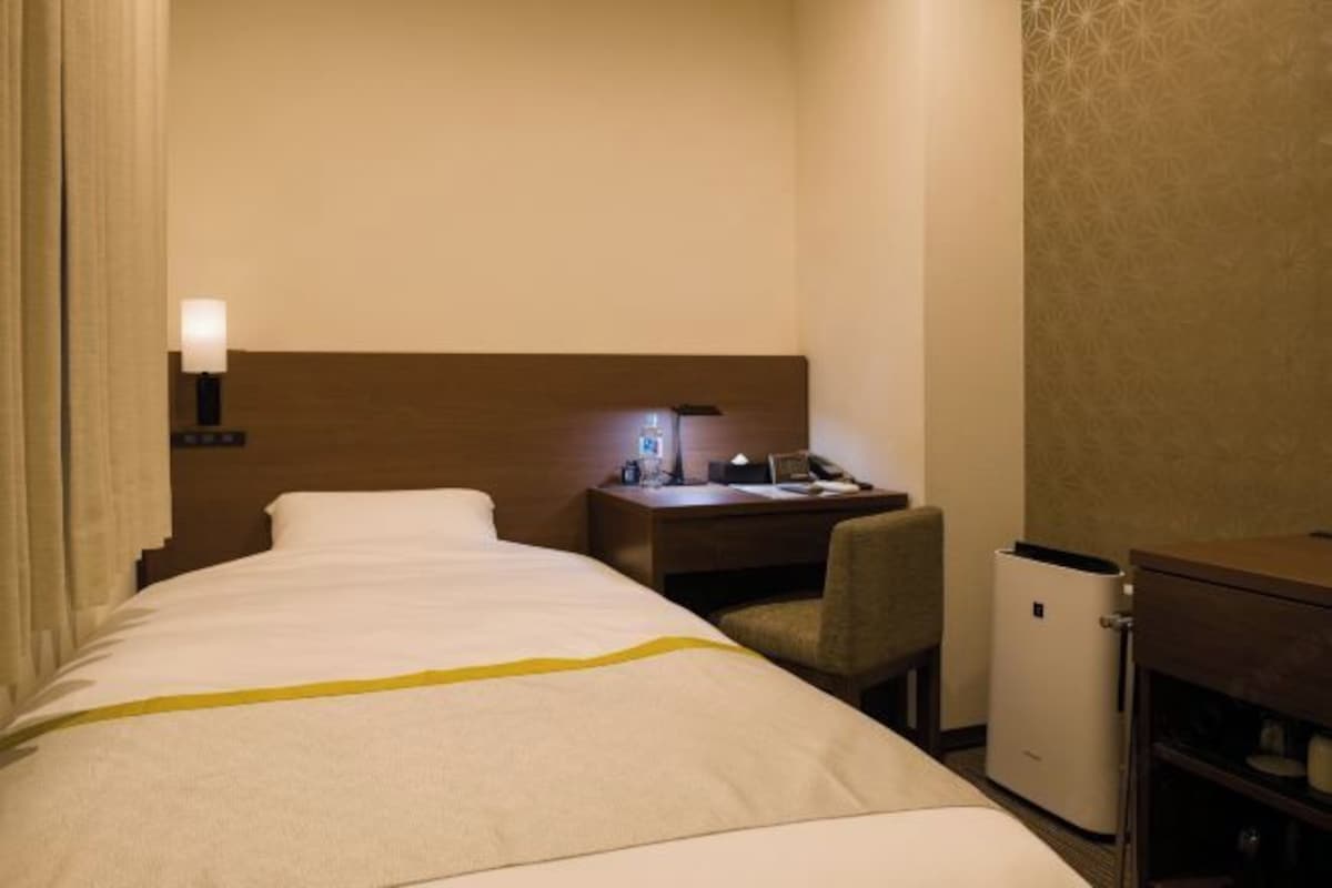 Quarto de hotel com cama, escrivaninha com luminária, cadeira e armário pequeno.