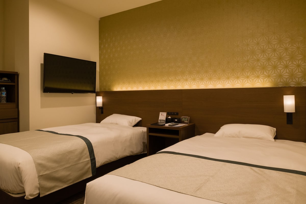 A hotel room with two beds, a flat-screen TV mounted on the wall, and a nightstand with a lamp and a small table.