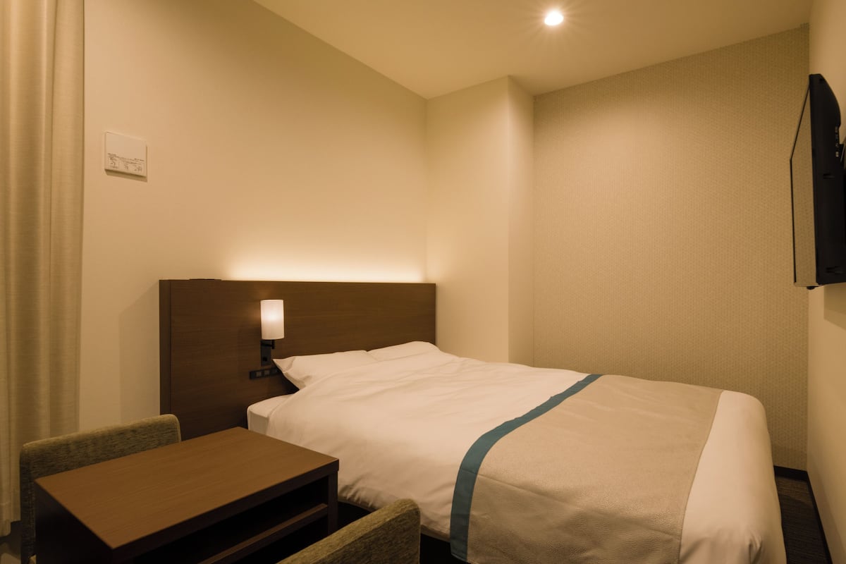 A hotel room with a single bed, a bedside table, a chair, and a flat-screen TV mounted on the wall.
