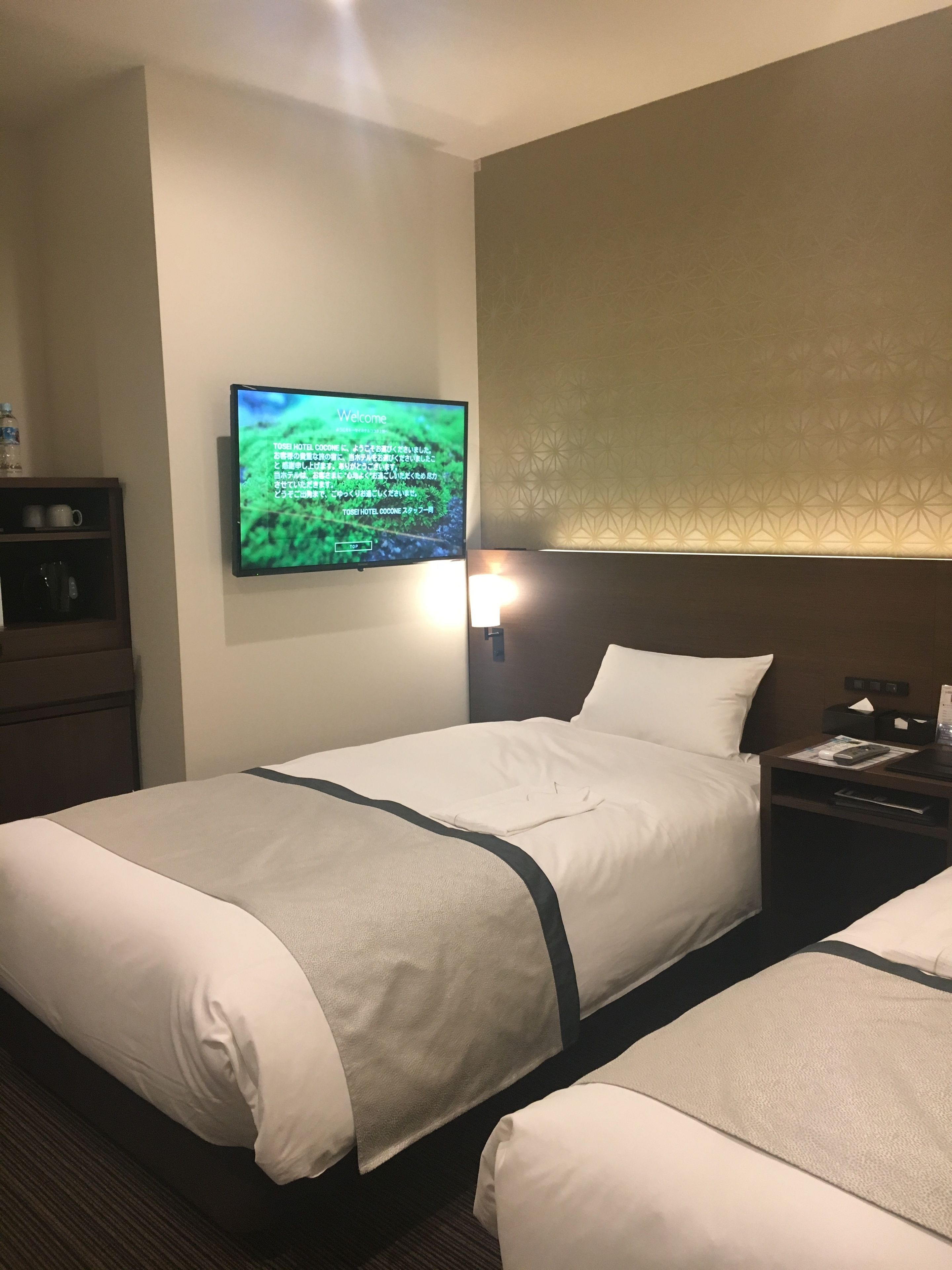 A hotel room with two beds, a TV on the wall, a nightstand, and a shelf with items.