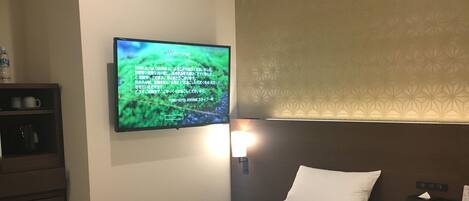 A hotel room with two beds, a TV on the wall, a nightstand, and a shelf with items.