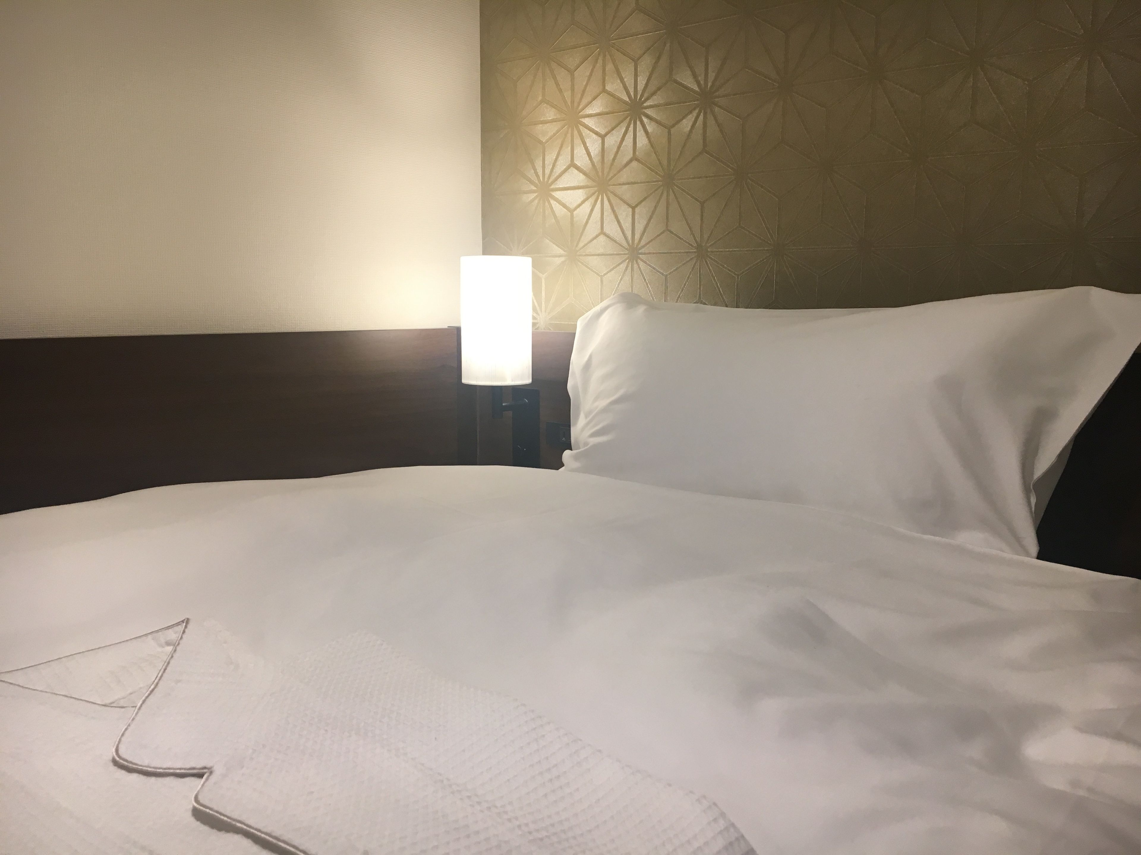 Una cama bien hecha con sábanas blancas y un cojín, una lámpara de noche y un papel tapiz con estampado geométrico.