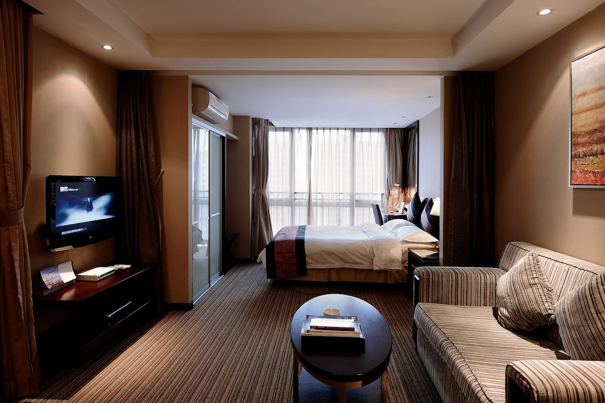 A hotel room with a bed, a sofa, a TV, a desk, and a large window.