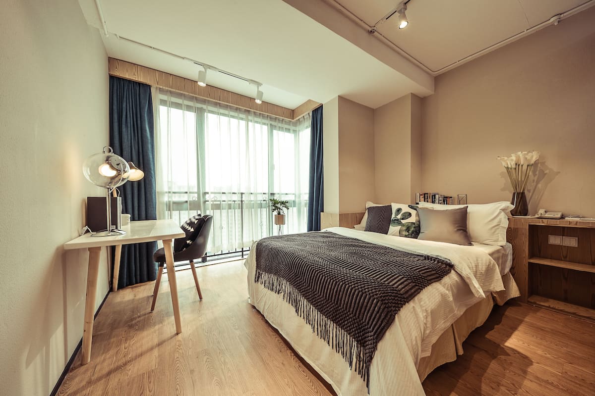 A modern hotel room with a wooden floor, a desk with a lamp, a bed with a patterned blanket, and a nightstand with a vase of flowers.