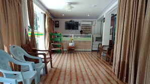 A room with a patterned wooden floor, plastic chairs, a wooden table, and a counter with a TV above it.