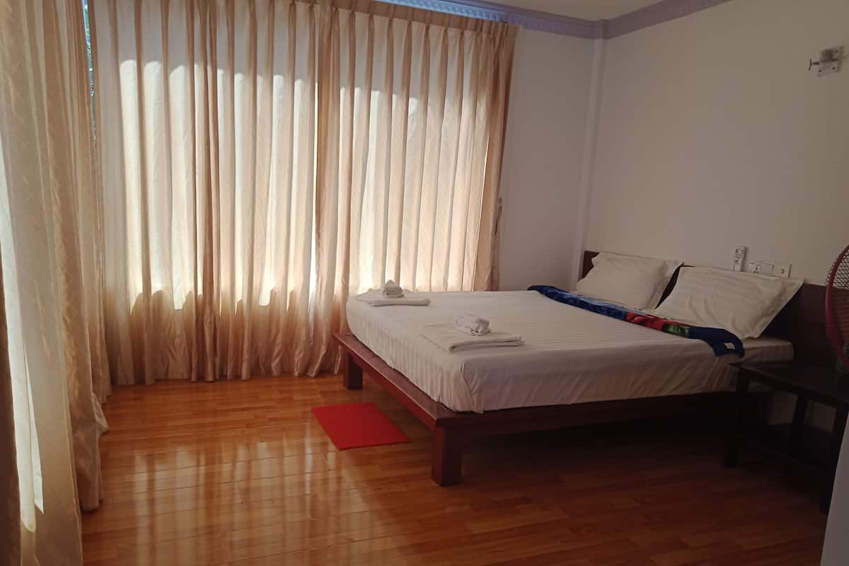 Une chambre avec un plancher en bois, un lit recouvert de draps blancs, un tapis rouge et une fenêtre ornée de rideaux légers.