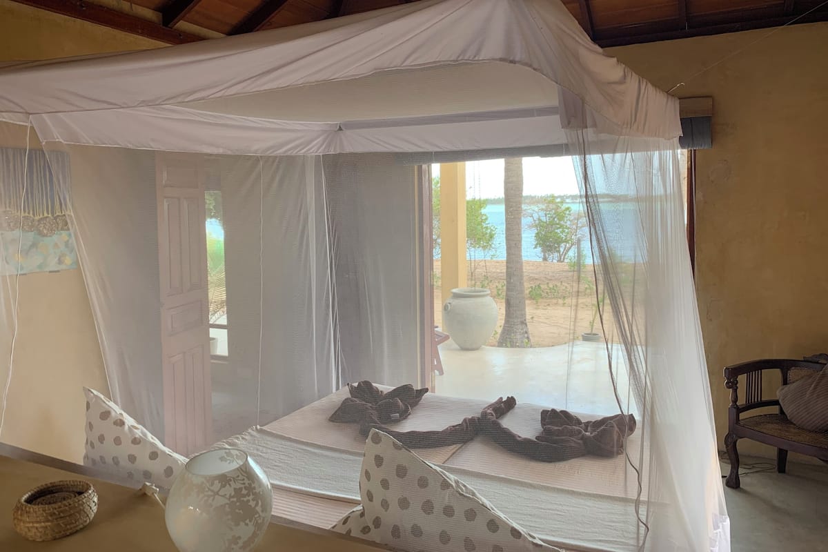 Una habitación con una cama bajo un dosel blanco, una ventana con vista a la playa y un banco de madera.