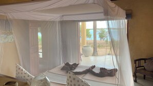 Una habitación con una cama bajo un dosel blanco, una ventana con vista a la playa y un banco de madera.