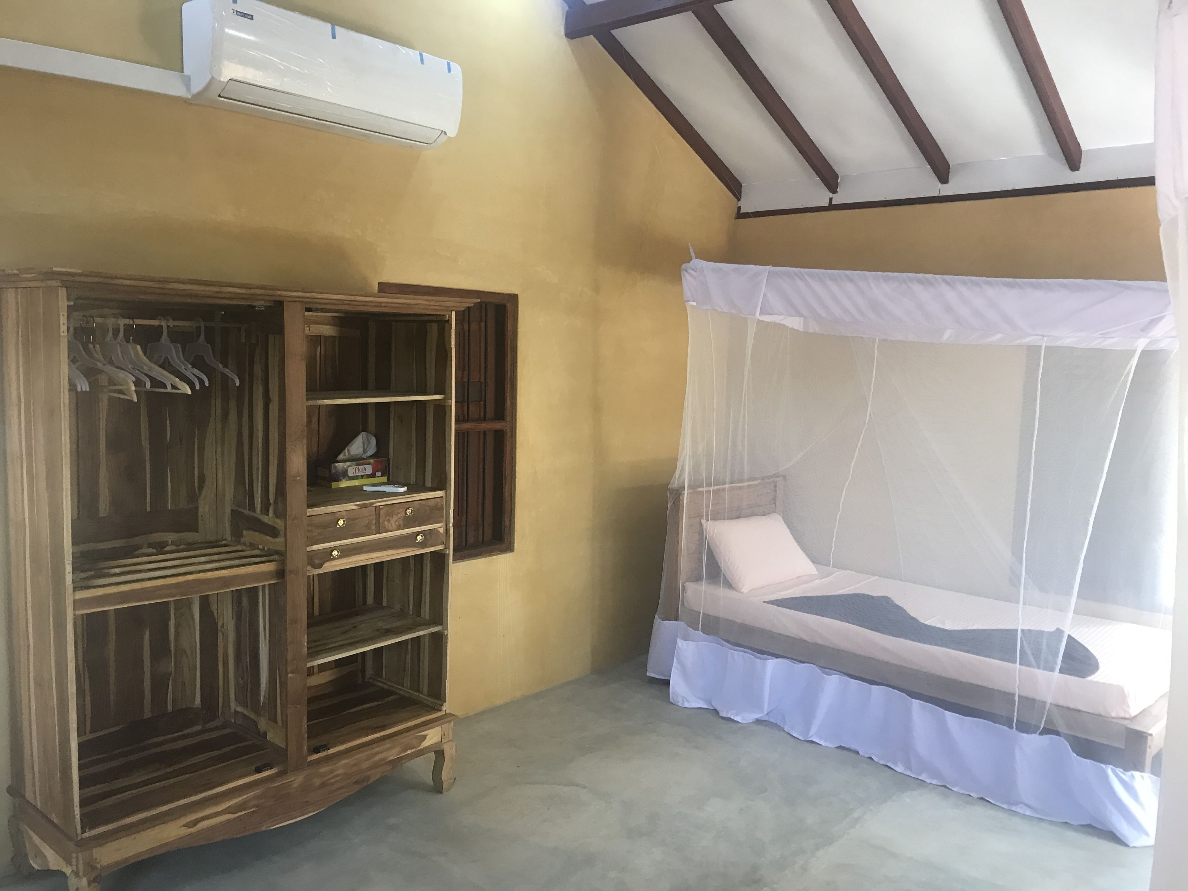 A room with a wooden wardrobe, a bed with a mosquito net, and an air conditioning unit on the wall.